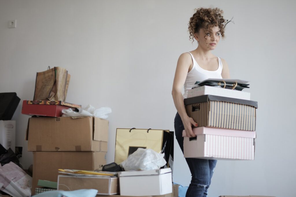 a woman carrying cardboard boxes outside of a room with clutter